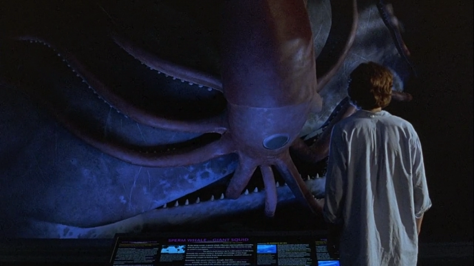 Walt, in a blue shirt, looks towards the diorama of the squid and the whale. He is small on the screen, the huge size of the two creatures dwarfing him.